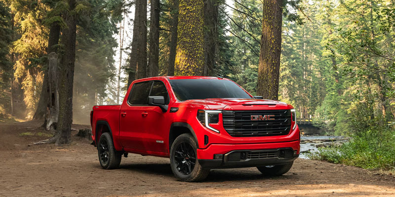 Red 2026 GMC Sierra 1500 pickup truck parked on a dirt path in a lush evergreen forest.
